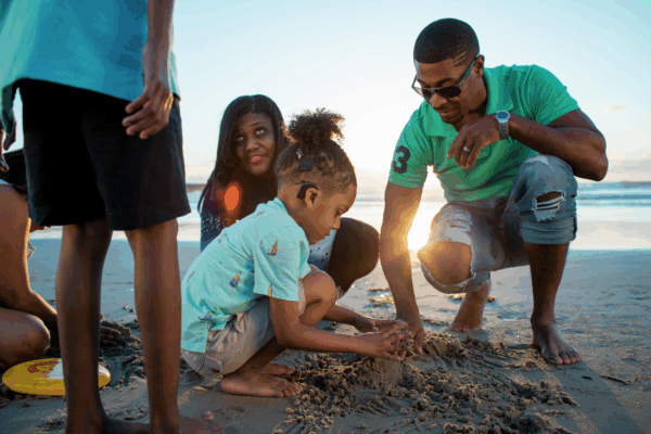 A family plays on a sandy beach during sunset. A young child with curly hair and a Nucleus 8 Sound Processor is crouched down, playing with the sand. An adult male and female kneel next to the child.