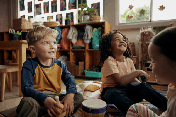 Children sitting on a rug in a classroom, playing musical instruments and smiling, with shelves and artwork in the background.