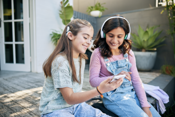 Two girls smiling and looking at a smartphone outdoors; one wearing a Nucleus 8 Sound Processor and the other wearing headphones.