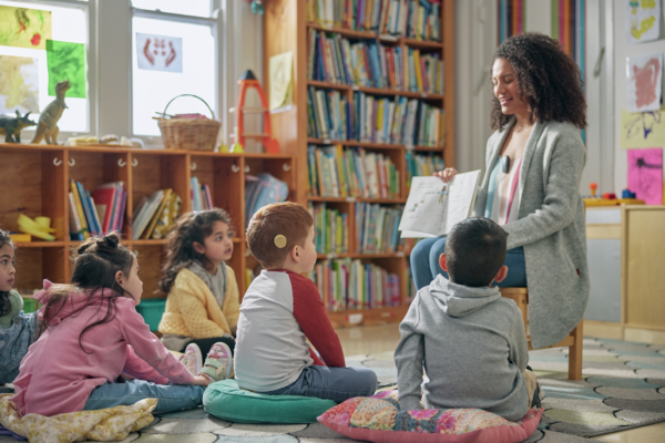 A teacher reads a book to children sitting on cushions in a classroom. A red-haired boy sitting in the front row wears a Kanso 2 Sound Processor.