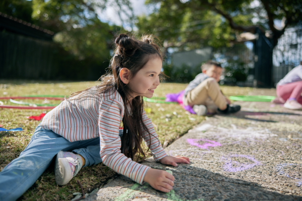 A young girl with a cochlear implant visbile, with long dark hair, wearing a striped long-sleeve shirt and jeans, is joyfully drawing on the pavement with colorful chalk. She is sitting on the ground with a bright smile on her face, engaged in creative play.