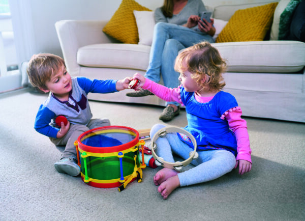Two toddlers playing with musical toys on a carpeted floor in a living room, with a woman sitting on a sofa in the background.