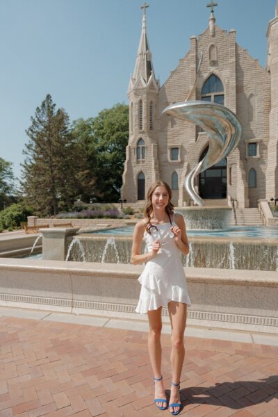 Addy, a girl with single-sided deafness who uses the Osia System, posing for a photo, standing in front of a fountain on her campus. She is holding a stethoscope around her neck.