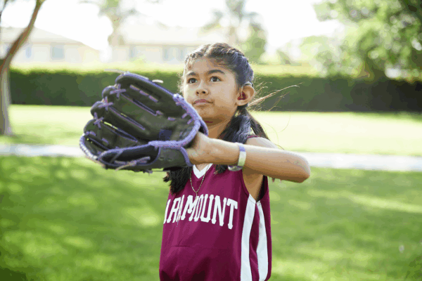 A young girl in a maroon sports jersey, wearing a hearing device, holds up a black baseball glove outdoors on a sunny day.