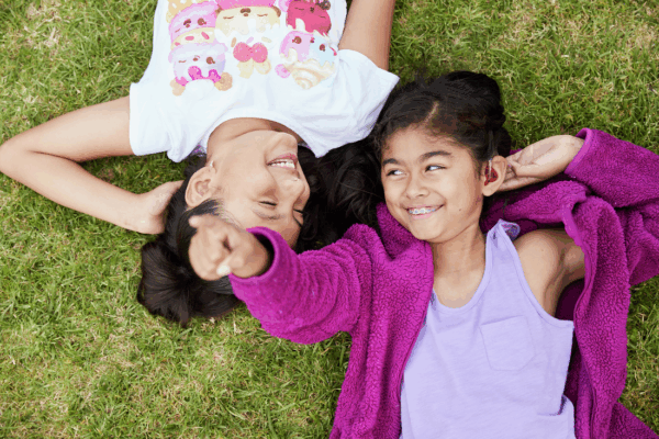 Two children lying on green grass, smiling and pointing, one wearing a white t-shirt, the other in a lavender shirt and purple jacket, touching her cochlear implant sound processor behind her ear.
