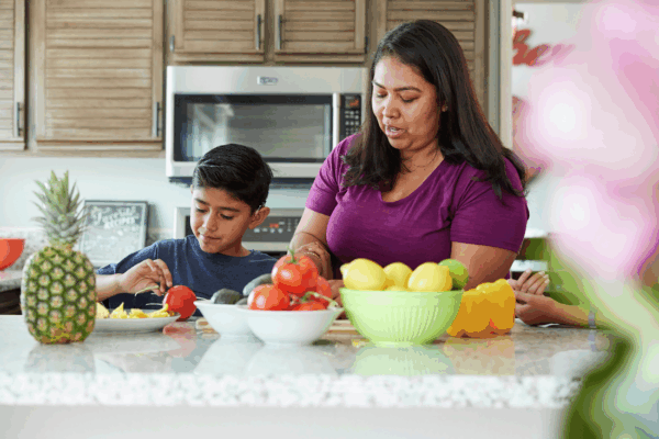 A woman and a boy, wearing a cochlear implant, preparing food in a kitchen with fresh fruit and vegetables on the counter.