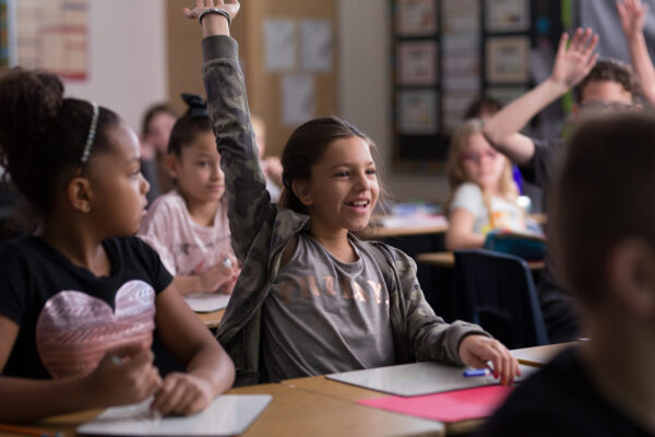 The image depicts a lively classroom setting with several children sitting at desks. The focus is on a student in the center, a young girl with her hand raised enthusiastically.