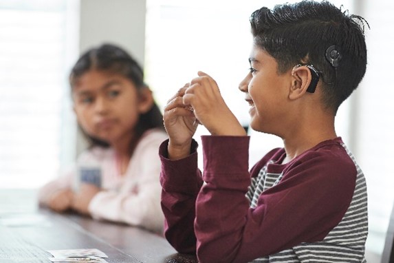 A smiling boy wearing Nucleus 8 Sound Processor sits at a table with a girl who is watching him.