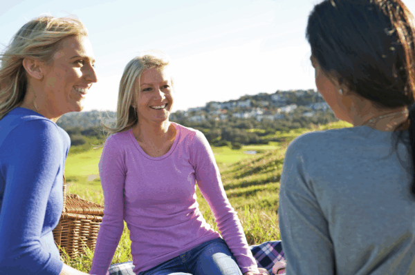 Three women, on a sunny day, smiling and sitting on a plaid blanket in a field with a picnic basket