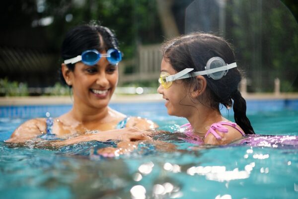 Mother and daughter are in a pool together, wearing goggles and smiling. The young girl is wearing the Aqua+.