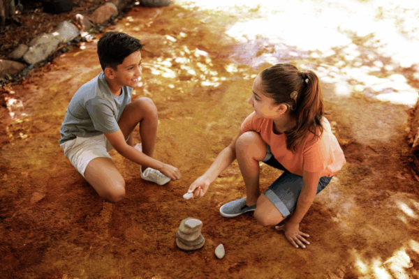 The image shows two children engaging in play on a sunlit patch of earthy ground, one with a Cochlear Nucleus 8 Sound Processor. They are both kneeling, facing each other, amidst shadows cast by nearby foliage.
