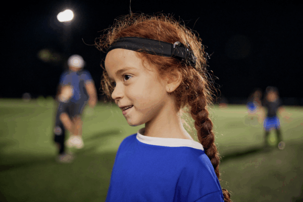 A young child with red braided hair, wearing a blue sports jersey and a black headband with sound processor, stands on a lit sports field at night.