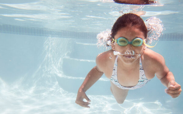 A girl wearing goggles, swimming underwater in a pool.