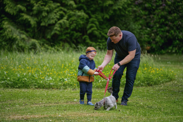 A man with a small gray dog on a leash hands the leash to a young boy. The boy is wearing a Baha® SoundBand®.