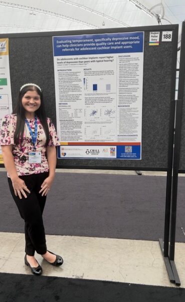 Skylynn stands next to a piece of her graduate work as an audiologist and shares how she gained confidence with her cochlear implant.