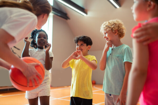 Four excited children are in a gym, gathered around a woman who is holding a basketball.