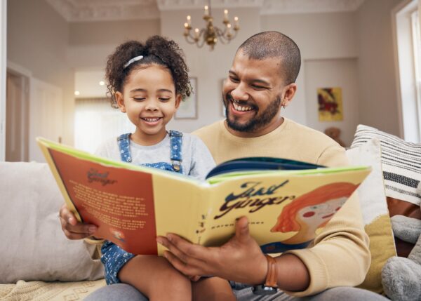 A young girl sits on a man’s lap on a couch. They read together.