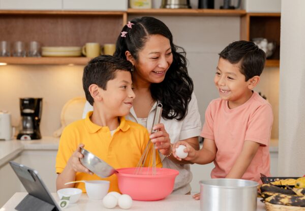 A woman stands in a kitchen with two young boys. They are helping her bake by adding ingredients into a bowl.