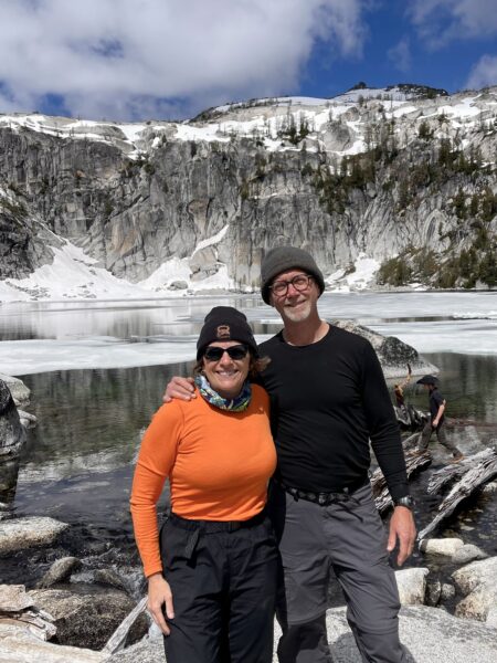 Hank and Kathy, who won back their shared adventures as a couple, after Kathy's hearing loss, smiling and standing in front of a snow covered cliff, in what may be Alaska. 