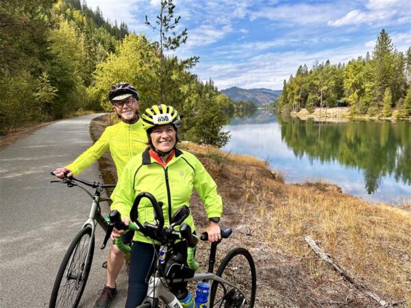 Hank and Kathy, who won back their shared adventures as a couple, with Kathy's cochlear implant, posing in high visibility jackets, next to their mountain bikes, in front of a bike path and beautiful backdrop of a river, mountains and forestry. 