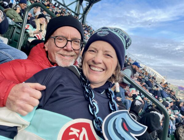 Hank and Kathy, who won back shared adventures as a couple, after Kathy's cochlear implant, smiling for a photo while attending a Seattle Kraken outdoor hockey game. Kathy is wearing a Kraken chain necklace, jersey and beanie. 