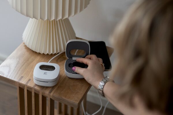 A hand is placing a Cochlear Kanso 3 Sound Processor in a Home Charger on a bedside table.