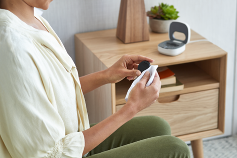 A woman wipes off a Cochlear Kanso 3 Sound Processor with a soft white cloth. A Kanso 3 Home Charger is visible on a nightstand