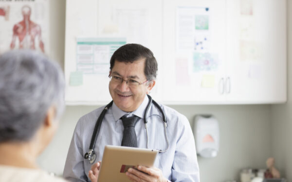 Pictured is a male doctor, middle-aged that is smiling at a patient and holding an iPad in his hands. 