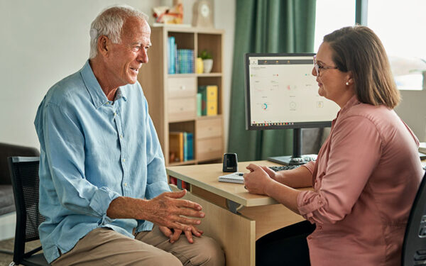 An older man with white hair and a Kanso Sound Processor on his head, speaking to his hearing health professional and smiling at the conversation. 