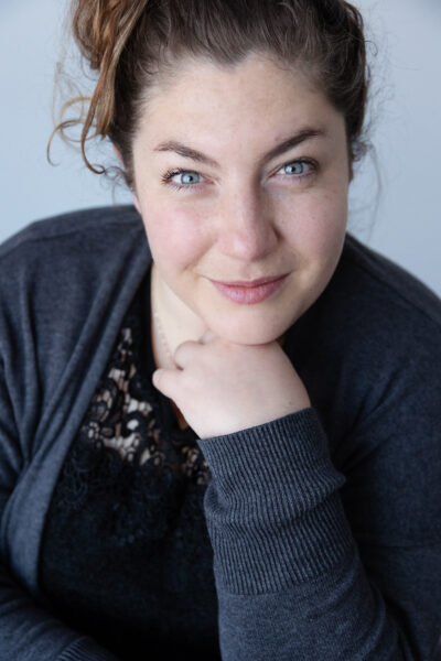 Kim, whose cochlear implant journey is featured, pictured as an adult with a professional headshot photo, where she is smiling with her chin resting on her hand, while it holds her head up.