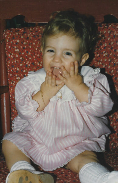 Kim, whose cochlear implant journey is featured, shown as a small child, holding her hands to her face in a smile for the camera.