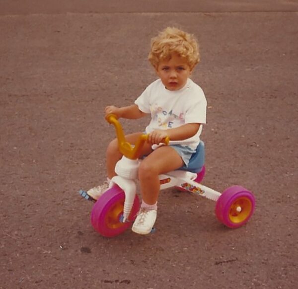 Kim, whose cochlear implant journey is featured, shown as a small child, riding on a white and pink toy tricycle. 