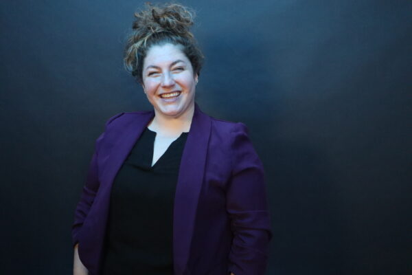 Kim, whose cochlear implant journey is featured, pictured as an adult with a professional headshot photo, where she is smiling in a purple blazer against a dark backdrop.