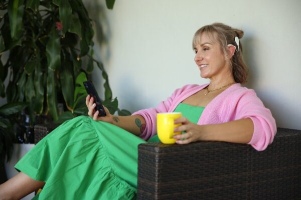 A recipient looking at her mobile phone showing her white Nucleus 8 Sound Processor, smiling and enjoying a cup of coffee; Ready-to-Wear for Cochlear Nucleus sound processor upgrades.