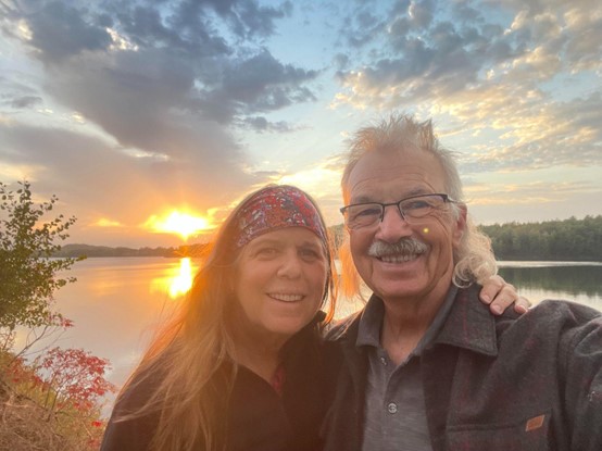 Tracy, who received clarity from her cochlear implant, pictured in front of a lake at sunset, smiling with her husband.