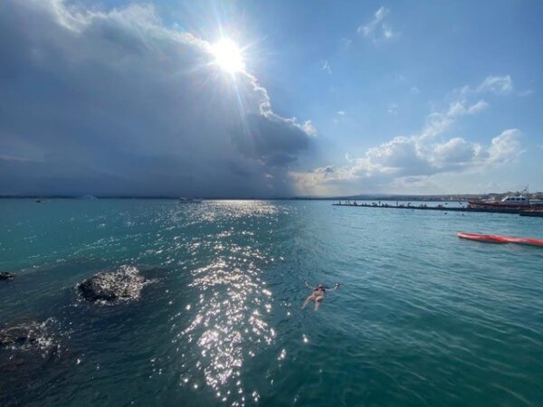 Linda, who received better hearing from her cochlear implant, pictured floating on her back in the sea, completely surrounded by blue water. The sun is peaking through the clouds above.