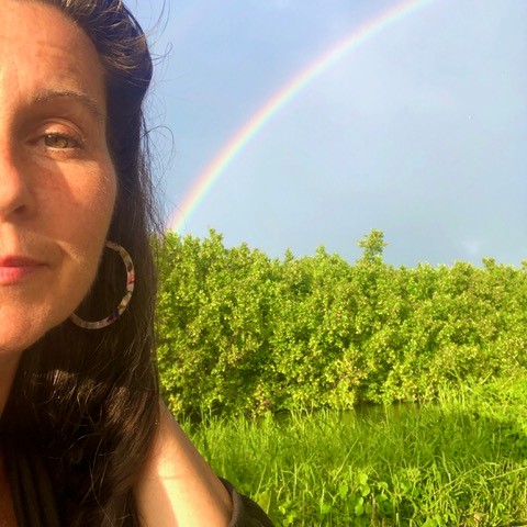 Linda, who received better hearing from her cochlear implant, smiling in a large green field filled with trees and wild grass, with a rainbow in the background.