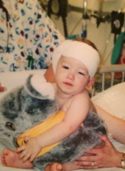 Broc, the 2025 Hear-o of the Year, as a baby sitting in a crib with a bandage around his head after his cochlear implant surgery.