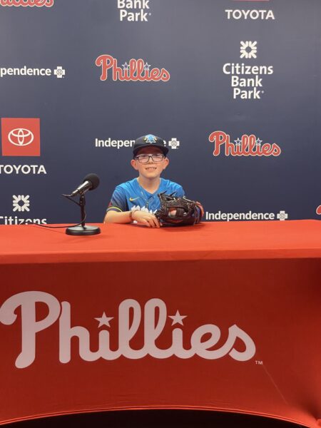 Anthony, a young boy with single-sided deafness and a cochlear implant, smiling for a picture, dressed as a baseball player, sitting at the Philadelphia Phillies media table.