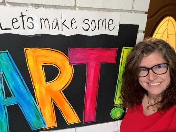 photo of woman with glasses standing with a sign that says “let’s make some art”; inspire us recipient artwork blog