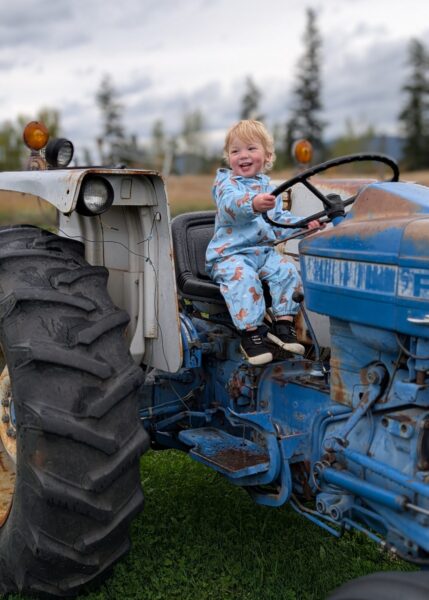 Lawson, a toddler who received a cochlear implant, smiling while sitting on a blue tractor in a wooded field.