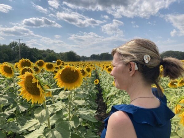 A picture of Shanna, who received a cochlear implant due to progressive hearing loss, standing in a field of sunflowers on a cloudy day. Her sound processor is visible behind her ear.