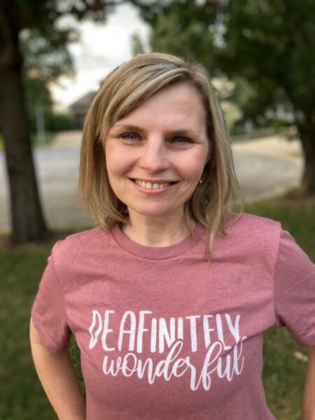 A picture of Shanna, who received a cochlear implant due to progressive hearing loss, smiling and wearing a “Deafinitely wonderful” t-shirt showcasing her podcast of the same name. 