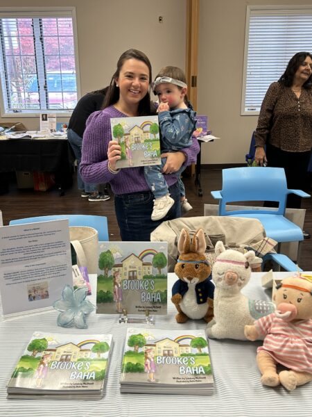 Brooke, a young girl born with microtia & atresia, who uses the Baha Start System, being held by her smiling mother Lindsey. They are holding a copy of the book ‘Brooke’s BAHA.’