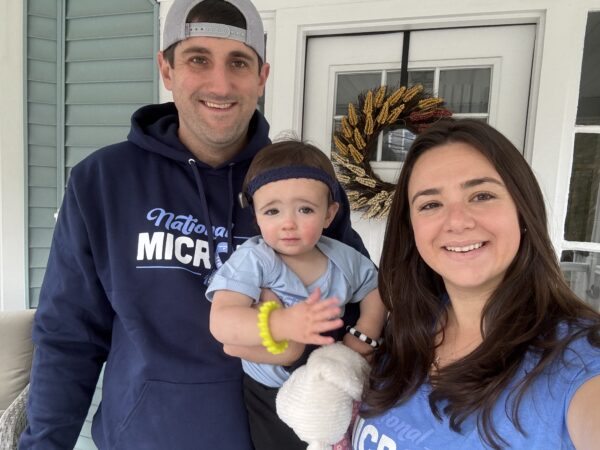 Brooke, a young girl born with microtia & atresia, who uses the Baha Start solution, being held by her mother Lindsey, and her father Ryan, on the porch of a sage green home, smiling for a family photo.