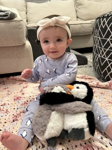 Brooke, a young girl born with microtia & atresia, pictured wearing the Baha Start solution with a Baha Softband around her head, smiling at the camera, sitting on the floor with penguin stuffed animals.