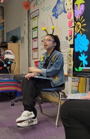 Amelia, who received early intervention after a bilateral sensorineural hearing loss diagnosis, reading the book she authored while sitting in front of a classroom with a chalkboard and pictures behind her.