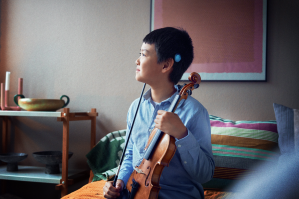 A profile photo of a pre-teen boy, where the light blue Osia 3 Sound Processor is shown above his ear. He is holding a violin and smiling as he looks to the side. 