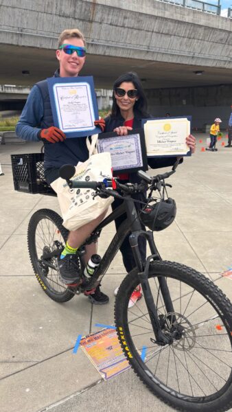 Pictured is Mickey, who was raised as a child with hearing loss, as an adult sitting on a mountain bike, holding certificates and smiling with his mother, Michelle who is standing next to him.