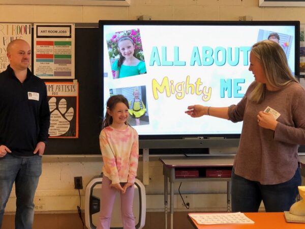 Mila smiling and presenting to her class with her Mom and Dad.